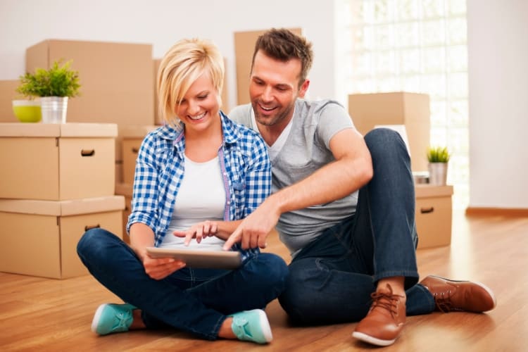 Man and woman sitting on the floor in a room looking at a tablet while smiling with moving boxes behind them.